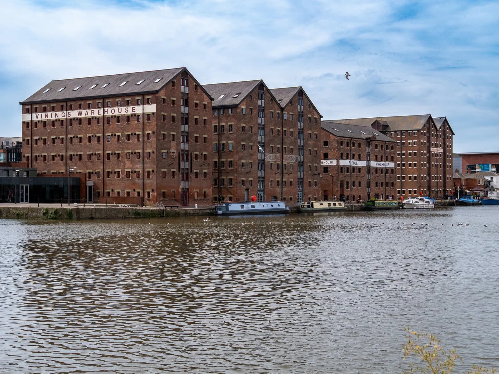 Several red brick warehouse buildings with many windows line a calm river, under a partly cloudy sky.